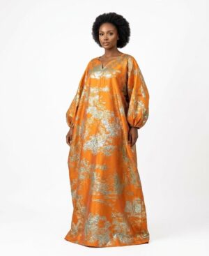 Woman wearing a voluminous orange and metallic silver kaftan dress with bishop sleeves against a white background.