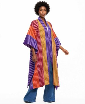 Full-length view of a woman wearing a long-line purple and orange striped African woven kimono jacket over blue jeans.
