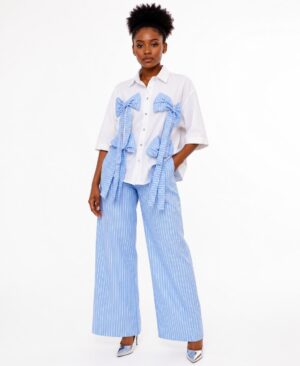 Woman wearing a white short-sleeved shirt with four large blue pinstripe bows and matching blue and white striped wide-leg trousers.