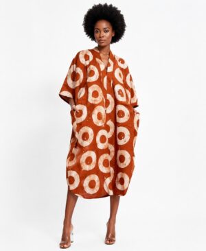 A Black woman with an afro hairstyle wearing an oversized burnt orange and cream batik print kaftan dress with side pockets.