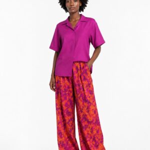 Black woman wearing a fuchsia short-sleeve shirt and orange-fuchsia batik print wide-leg trousers against a white background.
