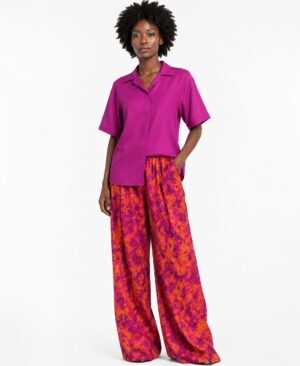 Black woman wearing a fuchsia short-sleeve shirt and orange-fuchsia batik print wide-leg trousers against a white background.
